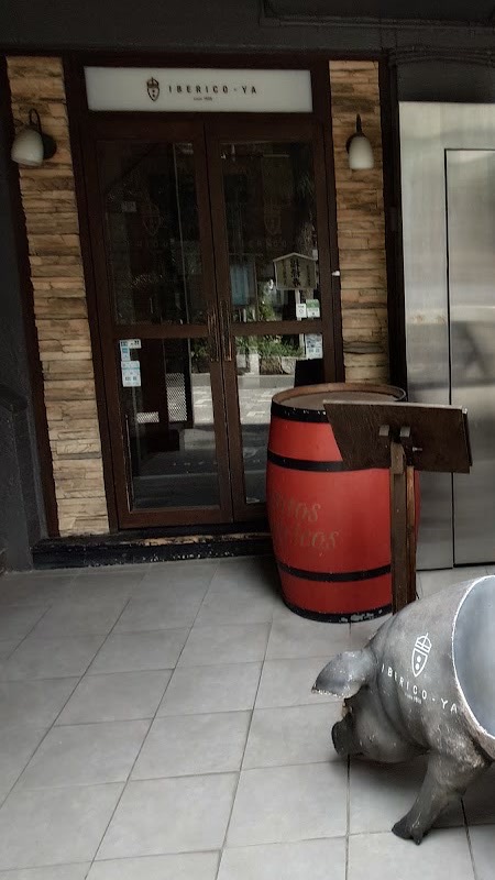 建物のエントランス内にある入口。紋章付きの白い「IBERICO-YA」のサイン、濃い木枠のガラス扉、積み石の柱と二つの壁灯、赤い樽、脇腹に紋章の入ったグレーの豚の置物。白いタイルの床。