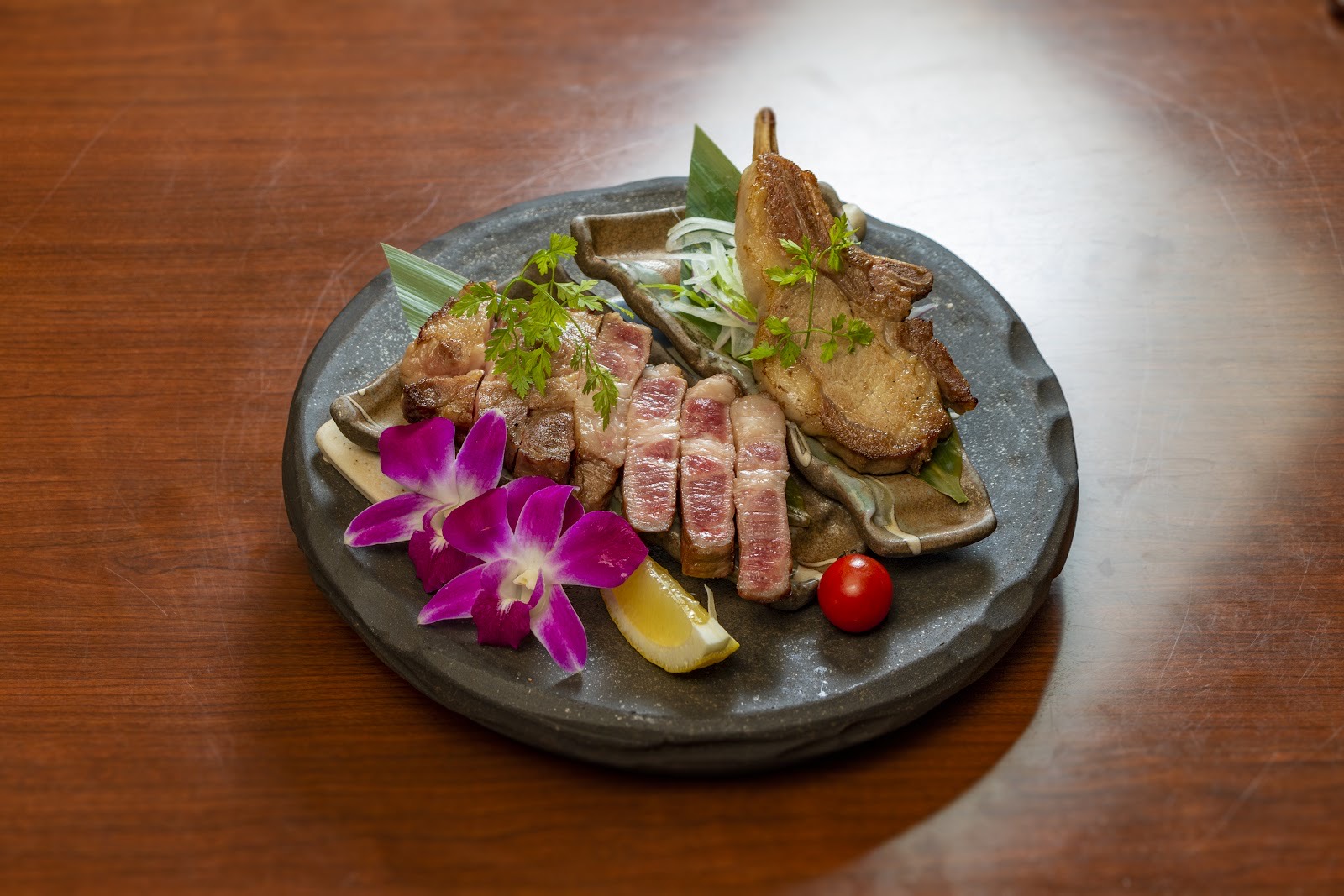木のテーブルに置かれた丸い灰色の石皿。スライスした骨付きの豚肉、紫の蘭、レモン、ミニトマト。