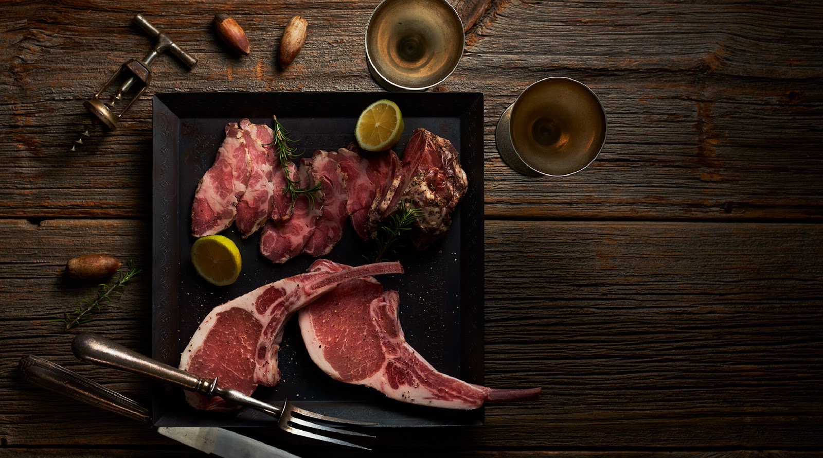 古木の上に置いた黒い板。生の骨付き豚肉と切り身、どんぐり、レモン、コルク抜き、グラス二つ。真上から。