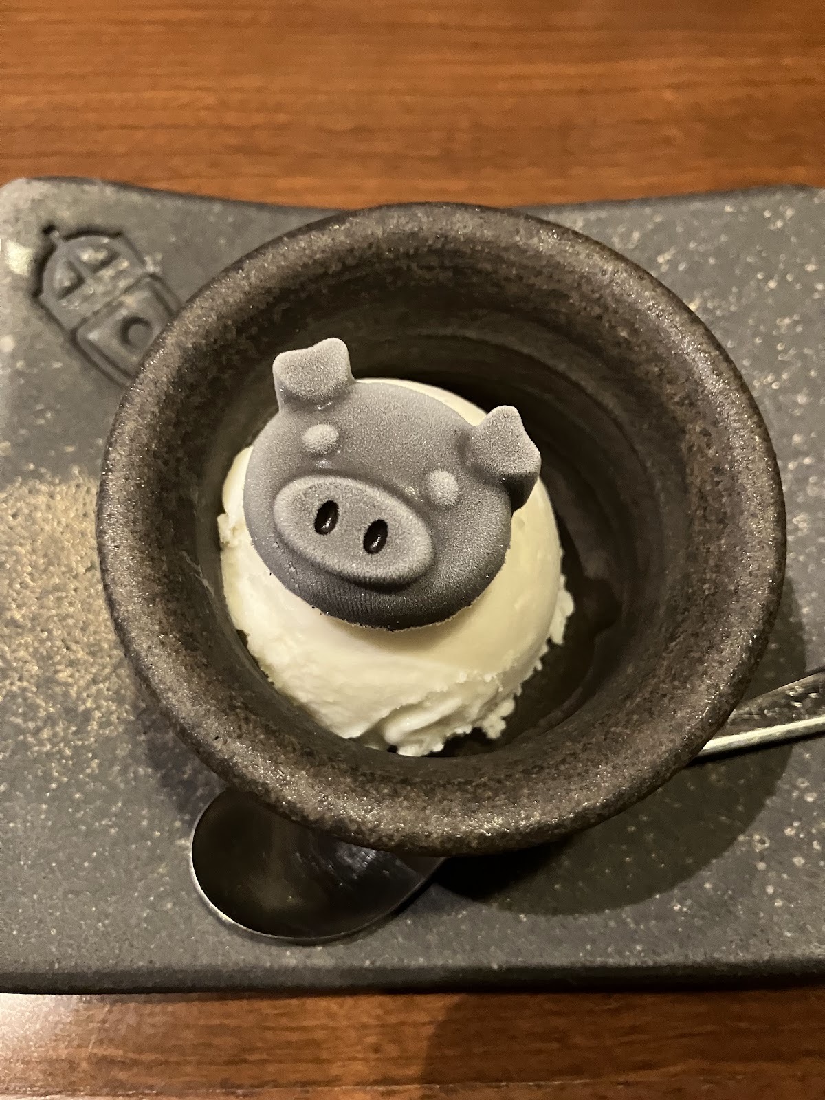 黒い石の器に入ったデザート。白いクリームの上に灰色の豚の顔。横にスプーン。