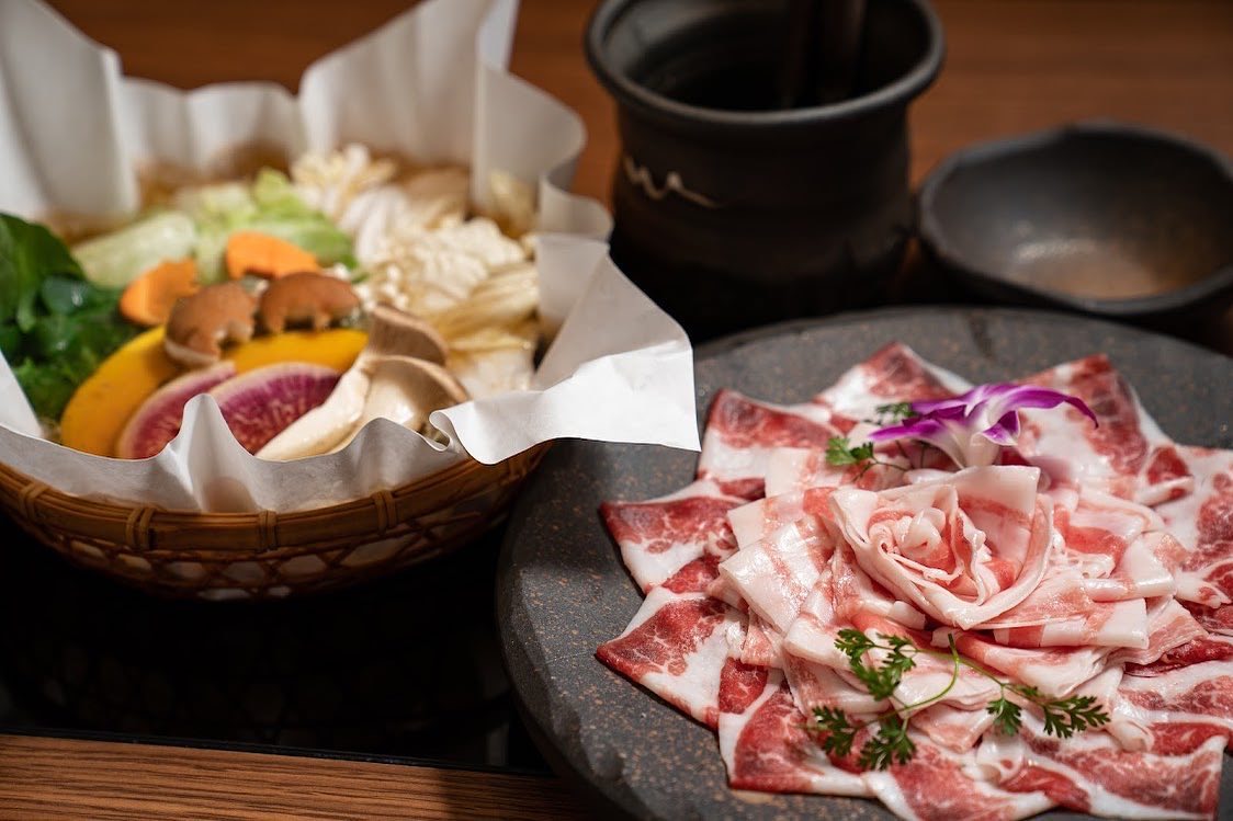 鍋のセット。灰色の石皿にバラの形に盛った薄切りの豚肉と蘭、野菜のかご、黒い鍋と器。